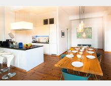 Moderne Wohnküche mit weißer Einbauküche, zentraler Kücheninsel mit Barhockern und hölzernem Esstisch in heller Umgebung. Über dem Tisch hängen mehrere Glühbirnenlampen, und an den Wänden sind ein Stadtplan und andere Dekorationen angebracht.