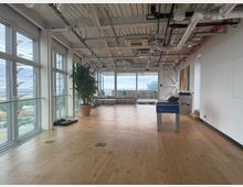 Open-plan loft interior with wood floors, floor-to-ceiling windows on two sides, and an exposed ceiling with visible ducts, pipes, and lighting. A potted tree, low sectional seating near the windows, and a foosball table sit in the spacious room with a city view beyond.
