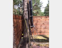 Eine Outdoor-Dusche, die an einem Baum befestigt ist, mit einem schwarzen Solarpanel, Duschkopf und Wasseranschlüssen. Die Umgebung ist naturbelassen, mit Holzwänden als Sichtschutz und Bäumen im Hintergrund.