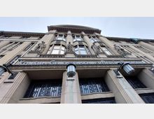 Fassade eines historischen Gebäudes mit der Aufschrift „Konsum-Genossenschaft Berlin“, verziert mit mehreren Statuen und großen Fenstern. Vor dem Eingang befinden sich schmiedeeiserne Gittertüren und zwei Laternen.