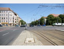 Kreuzung in einer städtischen Umgebung mit Straßenbahnschienen, Fahrbahnen für Autos und umliegender Bebauung. Links befinden sich mehrstöckige Wohnhäuser mit Geschäften im Erdgeschoss, während rechts Bäume und weitere Gebäude sichtbar sind. Oberleitungen für Straßenbahnen sind über der Kreuzung gespannt.