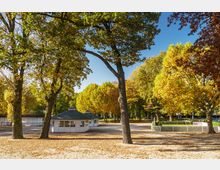 Parklandschaft im Herbst mit großen Laubbäumen, deren gelbe und orangefarbene Blätter den Boden bedecken. Im Hintergrund stehen weiße Pavillons mit grünen Dächern, umgeben von hellen Sandwegen.