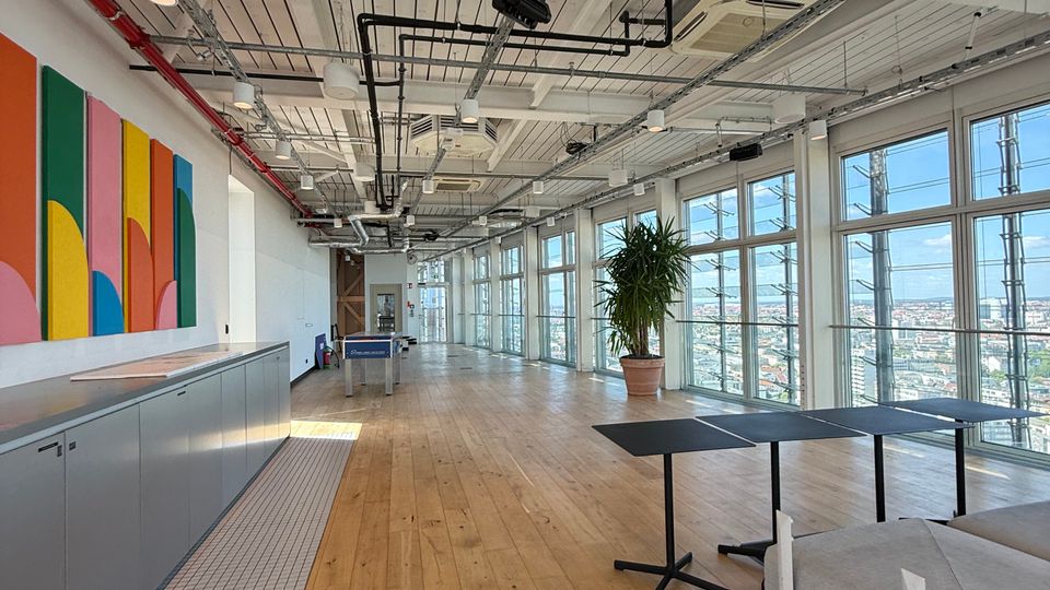 Open-plan modern office or studio with wood floors, an exposed white ceiling with pipes and track lighting, and floor-to-ceiling windows along one side overlooking a city. The space includes a long counter with colorful wall art, a potted plant, and scattered tables and lounge seating.