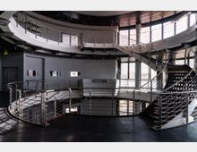 Offener, moderner Innenraum mit einer geschwungenen Treppe und umlaufenden Geländern aus Metall. Große Fenster mit Lamellen ermöglichen Lichteinfall, während die architektonische Gestaltung von Stahl und klaren Linien geprägt ist.