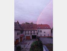 Courtyard of traditional residential buildings with red-tiled roofs, garage doors, and a central passageway, bordered by a paved area and small garden. A faint rainbow arcs across a pink evening sky above the rooftops.