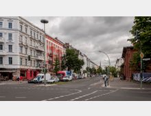 Straßenszene in einer Stadt mit mehrstöckigen Altbauten, teils in Weiß und Rot gestrichen, an der linken Seite. Die Straße ist von einer Baumreihe umgeben und zeigt geparkte Autos und Läden im Erdgeschoss der Gebäude. Rechts ist ein Backsteinbau mit Straßenschildern und Ampeln sichtbar, während der Himmel bewölkt ist.