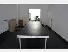 Ein schlichter Besprechungsraum mit einem langen schwarzen Tisch in der Mitte, umgeben von schwarzen Stühlen. Im Hintergrund führt eine Türöffnung zu einem helleren Raum mit Büromöbeln.