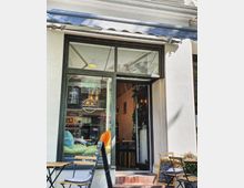 Fassade eines kleinen Cafés mit breitem Fenster, durch das bunte Sitzkissen sichtbar sind. Vor dem Eingang stehen eine schwarze Tafel mit Menüangeboten und mehrere kleine Tische und Stühle auf dem Gehweg.