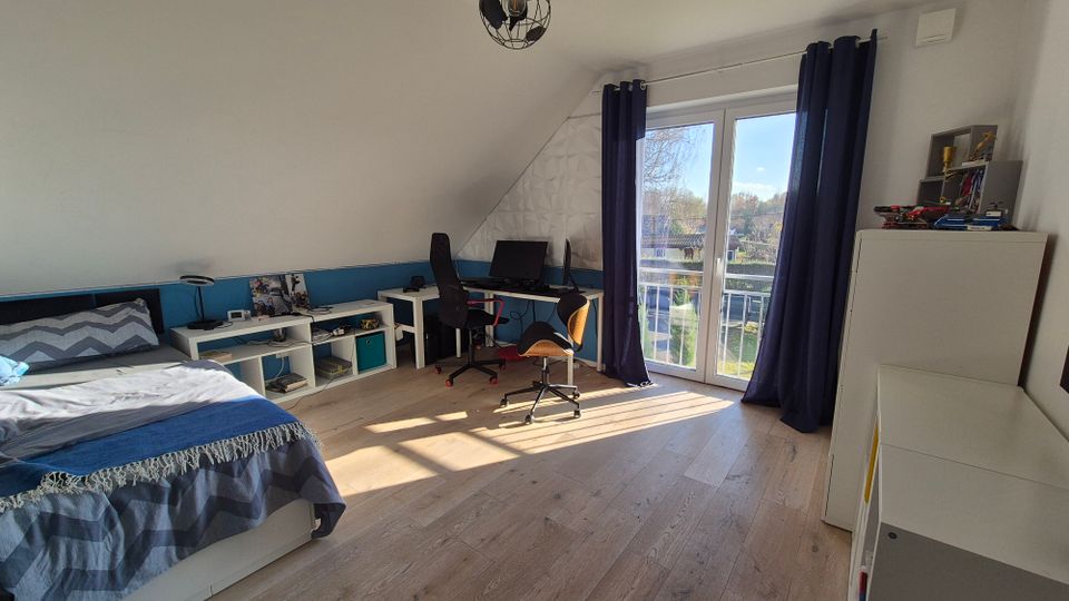 Ein modernes Schlafzimmer mit Dachschräge, einem Bett links, einem Schreibtisch mit Computer vor der Rückwand und einer weißen Balkontür mit blauen Vorhängen. Helle Holzböden und weiße Möbel dominieren den Raum, der von Tageslicht durchflutet wird.