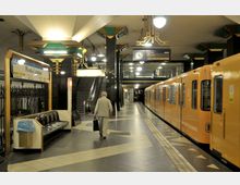 U-Bahn-Station mit gelbem U-Bahn-Zug auf der rechten Seite, Sitzbänken und einem Wegweiser auf der linken Seite. Die Station hat eine schwarz-goldene Art-Déco-Decke, eine Treppe und Rolltreppe im Hintergrund sowie Uhren und Informationsanzeigen.