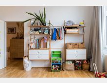 Kinderzimmer mit einer Holzregal-Kombination, bestehend aus weißen Schubladen, hängenden Kleidungsstücken, Körben und Deko-Objekten. Darunter befinden sich Aufbewahrungskörbe und Spielzeug, rechts ein Fenster mit einem bodenlangen Vorhang.