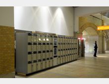 Reihe von Schließfächern in einem Bahnhof oder öffentlichen Gebäude, flankiert von gelben Klinkerwänden und Bögen. Ein Mann steht in der Nähe, und im Hintergrund sind Hinweisschilder und beleuchtete Bereiche zu sehen.