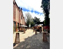 Ein gepflasterter Innenhof mit Backsteingebäuden auf der linken Seite, ausgestattet mit Holzbistrotischen und einigen Sitzbereichen mit Sonnenschirmen. Im Hintergrund stehen Verkaufsstände und hohe Bäume, die den Bereich umrahmen.