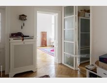 Wohnung mit hellem Holzboden und weißen Wänden; im Vordergrund eine weiße Heizkörperverkleidung mit abgelegten Gegenständen, daneben eine Glastür mit Milchglas. Im Hintergrund ein Zimmer mit farbigem Teppich, einem antiken Holzschrank und Tageslicht.