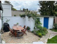 Ein kleiner, gepflasterter Hinterhof mit einem Holztisch und mehreren Stühlen in der Mitte, umgeben von Pflanzen und blühenden Sträuchern. Im Hintergrund steht eine weiß gestrichene Mauer mit einem Nebengebäude, dessen Tür in einem kräftigen Blau gestrichen ist, während im Vordergrund eine Rasenfläche sichtbar ist.