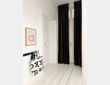 Bright, minimalist hallway with white walls and wide white plank flooring, featuring paneled doors and a tall doorway framed by long dark curtains. A small stained-glass transom window sits above the doorway, and a wall-mounted hook rack holds a large tote bag beneath a framed print.