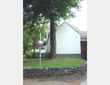 Ein kleiner Vorgarten mit einer niedrigen, aus Natursteinen gemauerten Mauer, einem großen Baum und einem weißen Gebäude mit Satteldach im Hintergrund. Ein Verkehrszeichen mit Halteverbot steht seitlich neben dem Baum.