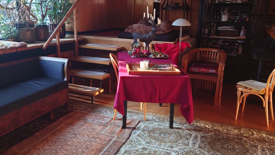 Wohnzimmer mit Holzwänden und gemütlichem Ambiente, ausgestattet mit einem orientalischen Teppich, einer roten Tischdecke auf einem Esstisch, Holzmöbeln und einer Sitzbank. Im Hintergrund befinden sich Pflanzen, Schränke, Regale und dekorative Elemente wie Kerzenhalter und Wandgemälde.