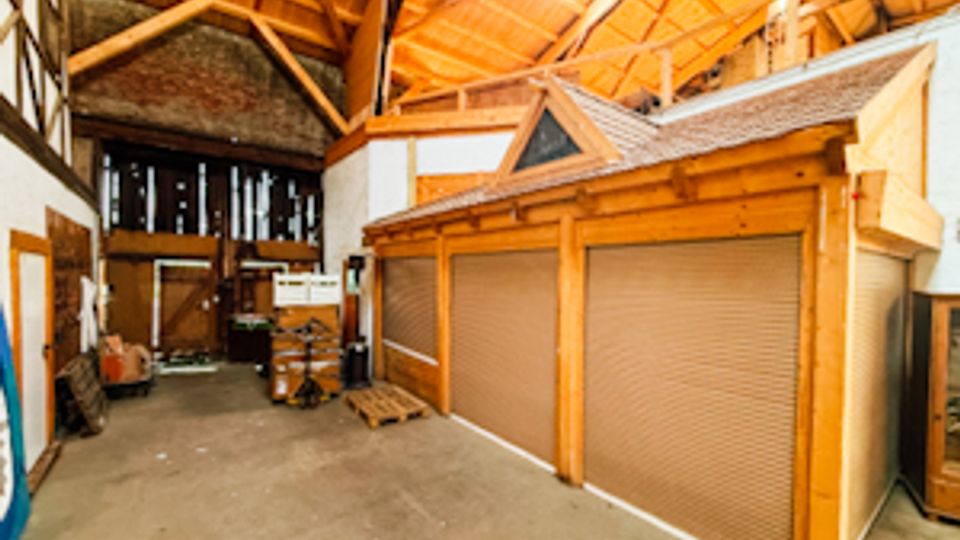 Innenansicht einer großen Lager- oder Werkhalle mit hoher Holzbinder-Dachkonstruktion und Oberlichtfenstern. In der Halle stehen eingebaute Holzboxen mit Rolltoren, daneben Betonboden sowie Regale und gelagerte Gegenstände entlang der Wände.