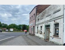 Straßenszene mit einem alten, stark renovierungsbedürftigen Gebäude auf der rechten Seite und einer belebten Straße daneben; im Hintergrund sind eine Grünfläche und geparkte Autos zu sehen, während ein Radfahrer auf dem Gehweg fährt.