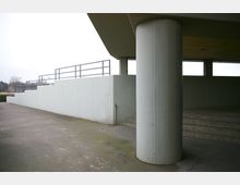 Außenbereich einer Betonkonstruktion mit einem großen, runden Pfeiler im Vordergrund, einer seitlich ansteigenden Rampe und mehreren Stufen rechts. Im Hintergrund sind Geländer und eine offene Umgebung mit Bäumen zu sehen.