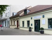 Straßenszene mit kleinen historischen Gebäuden, darunter ein cremefarbenes Haus mit Giebeldach und grünen Fenstern, das als Geschäft mit der Aufschrift "Home Dress" genutzt wird. Links daneben ein weiteres Gebäude mit aufwendiger Fassadengestaltung und roter Eingangstür, vor denen ein Gehweg mit einer Person entlangführt.