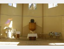 Innenraum einer Kirche mit schlichten, hellen Wänden. Im Zentrum steht eine Holzkanzel, darunter Tische mit weißen Decken, Stühlen und Schautafeln an den Seitenwänden. Oben sind drei Kirchenfenster mit Gittermuster zu sehen.