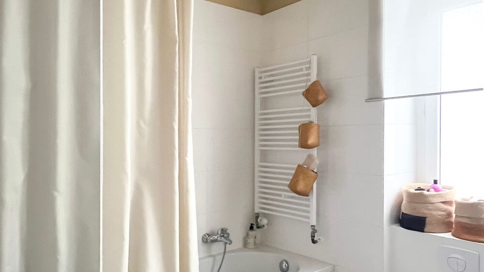 Bright bathroom with a white-tiled bathtub and shower curtain, a wall-mounted towel radiator, and a floating toilet beside a window ledge with storage baskets. Dark flooring contrasts with the white fixtures, and the ceiling is painted a warm beige.
