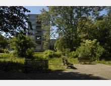Wohnblock mit sechs Stockwerken im Hintergrund, umgeben von grünen Sträuchern und Bäumen. Im Vordergrund stehen mehrere Holzbänke auf einem gepflasterten Platz, teils im Schatten der Vegetation.