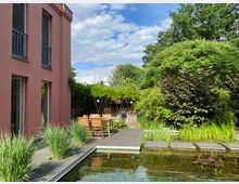Moderner Gartenhof neben einem rosafarbenen Gebäude mit großer Terrassenfläche, Holztisch und Stühlen unter einer begrünten Pergola. Im Vordergrund liegt ein rechteckiger Naturteich mit Seerosen, Schilfpflanzen und Trittplatten, umgeben von dichtem Baumbestand.