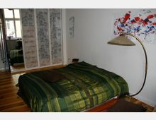 Schlafzimmer mit einem niedrigen Bett, das mit einer grün gestreiften Tagesdecke bedeckt ist. Im Hintergrund befindet sich ein Schrank mit weißen Schiebetüren und floralem Muster sowie ein großes Fenster, das Tageslicht hereinlässt. Ein gebogener Stehlampenarm und ein abstraktes Wandbild ergänzen den Raum.