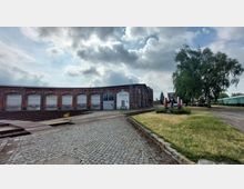 Backsteingebäude eines historischen Bahnbetriebswerks mit mehreren Toren, Eisenbahnschienen und einer Pflasterfläche im Vordergrund; rechts eine Grünfläche mit Baum und einem ausgestellten Radsatz.