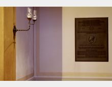 Wand in einem Innenraum mit einer Metall-Gedenktafel auf der rechten Seite, die eine Inschrift und das Relief eines Kopfes zeigt. Links befindet sich eine historische Wandlaterne mit zwei gläsernen Leuchtkörpern.