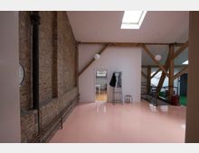 Ein Innenraum eines Lofts mit einer Kombination aus Sichtbacksteinwand und weiß gestrichenen Wänden. Sichtbare Holzbalken stützen die Decke, ein rosafarbener glänzender Boden und ein Dachfenster sorgen für Helligkeit, während eine offene Tür den Blick in einen angrenzenden Raum mit Tischen und Stühlen ermöglicht.