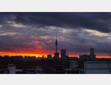 Städtische Skyline mit dem Berliner Fernsehturm im Zentrum, aufgenommen bei Sonnenuntergang. Der Himmel zeigt intensive Rot- und Orangetöne, die von dunklen Wolken überlagert werden.
