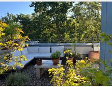 Grüne Dachterrasse mit einem Sitzbereich aus weiß gepolsterten Sofas, umgeben von Blumentöpfen mit Pflanzen und Bäumen. Im Vordergrund ein Tisch aus Stein, darauf kleine Pflanzgefäße; im Hintergrund Geländer und dichte Baumkronen.