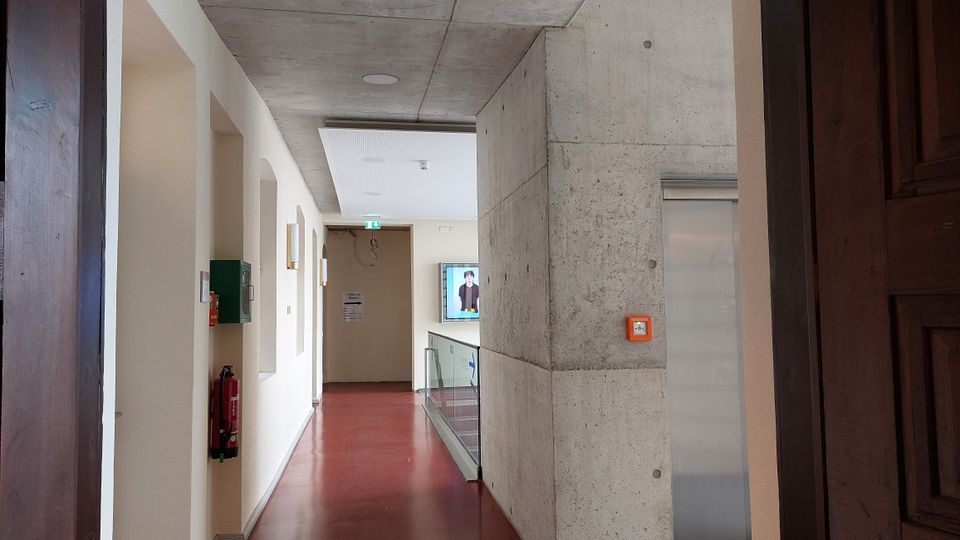 Korridor in einem modernen Gebäude mit Betonwänden, rotem Boden und einer abgehängten Decke. An der Wand befinden sich ein Feuerlöscher und eine grüne Box, während ein Bildschirm am Ende des Ganges sichtbar ist.