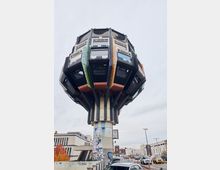 Futuristisches Gebäude mit einer kugelartigen Struktur auf einem schmalen, grafittibedeckten Pfeiler, das eine Mischung aus versetzten Fassaden und bunten Elementen zeigt; im Hintergrund eine urbane Straßenumgebung mit geparkten Autos und modernen Gebäuden.