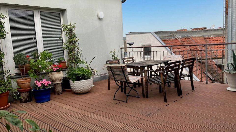 Rooftop terrace with wood decking, a metal railing, and a rectangular outdoor dining table surrounded by folding chairs. Potted plants and flowers line the wall near a large window, with views of neighboring red-tiled rooftops under a clear sky.