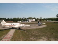 Kleiner Flugplatz mit zwei geparkten Kleinflugzeugen auf einer betonierten Fläche. Im Hintergrund stehen ein weißes Gebäude und weitere Flugzeuge auf einem weitläufigen Gelände, umgeben von Bäumen.