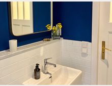 Kleines Badezimmer mit dunkelblauer Wand und weißen Fliesen. Ein rechteckiger Spiegel mit schmalem Rahmen hängt über einem weißen Waschbecken; daneben stehen eine Vase mit gelben Blumen und eine Seifenspenderflasche.