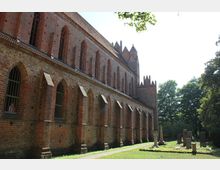 Backsteinbau einer historischen Kirche mit gotischen Spitzbogenfenstern, flankiert von einem gepflegten Außenbereich mit mehreren Grabsteinen. Im Hintergrund sind Bäume sichtbar, die die grüne Umgebung betonen.