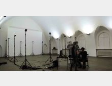 Ein großer, heller Raum mit gewölbter Decke und mehreren hohen, bogenförmigen Fenstern. Der Raum ist mit zahlreichen Stativlampen ausgestattet, und es stehen Tische sowie Stühle im Vordergrund, wo eine Szene mit mehreren Personen stattfindet.