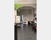 Ein moderner Tätowierladen mit dunklem Fliesenboden, einem großen Arbeitstisch in der Mitte und mehreren Tätowierstationen mit Stühlen und Liegen. An der Rückwand befinden sich gerahmte Illustrationen, ein kleiner weißer Schreibtisch und ein Heizkörper.