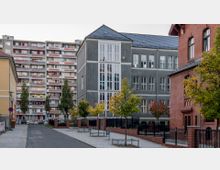 Straßenszene mit einem grauen Gebäude im Vordergrund, das große Fenster mit weißen Rahmen hat, flankiert von roten und gelben Backsteingebäuden. Im Hintergrund steht ein großes Wohnhaus mit zahlreichen Balkonen, umgeben von Bäumen und Gehwegen.
