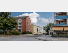 Eine städtische Straßenszene mit mehrstöckigen Wohngebäuden, darunter ein rotes Gebäude links und ein Gebäude mit Balkonen und blauen Geländern rechts. Im Vordergrund eine Kreuzung mit Kopfsteinpflaster, Verkehrszeichen und einem Schaufenster eines Fotogeschäfts.