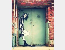 Abbildung einer verschlossenen, grauen Metalltür in einem Backsteinbau mit Graffiti-Kunst, das eine stilisierte Frau zeigt, die lässig an der Tür lehnt. Gerüstelemente und beschädigte Bereiche im Mauerwerk sind sichtbar.