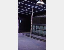 Innenraum eines Clubs oder Veranstaltungsortes mit dunklen Holzböden, einem diagonalen LED-Lichtmuster an der Decke, einer Metallstange mit Stickern und einer Wand mit mehreren alten Röhrenfernsehern. Im Hintergrund sind Gitterelemente zu sehen.