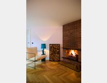 Modernes Wohnzimmer mit einem gemauerten Kamin mit brennendem Feuer, einer hölzernen Brennholzablage, einem Sessel mit hellem Stoffbezug und einer blauen Tischlampe auf einem Beistelltisch. Der Boden besteht aus Fischgrätenparkett, die Wände sind hell gestrichen.