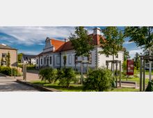 Ein kleines weißes Gebäude mit roten Ziegeldach und dekorativer Fassade, umgeben von einem gepflegten Garten mit Bäumen und Sträuchern. Im Vordergrund befindet sich ein gepflasterter Weg sowie eine Informationstafel.